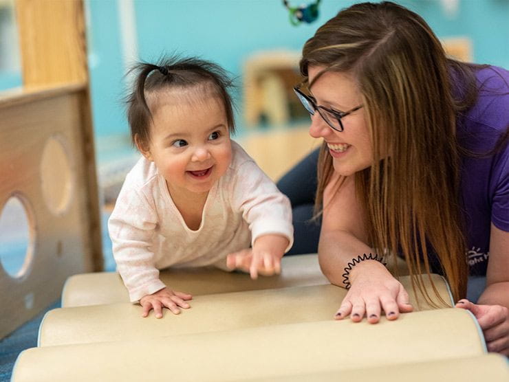 Little girl smiling and crawling on a mat next to a caregiver